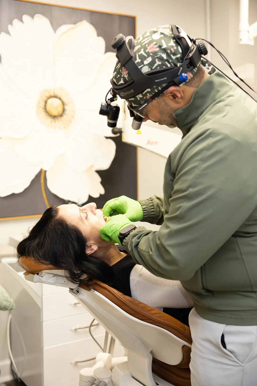 Dentist wearing loupes examining a patient in a brown dental chair under a floral painting — My Dental Hub in Skokie, IL