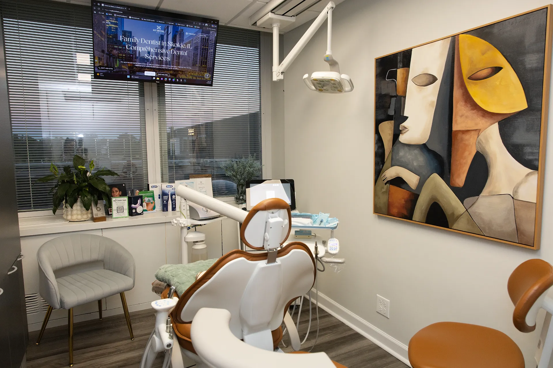 Dental operatory with white and brown chair, abstract artwork on wall, and a large TV showing the practice website — My Dental Hub in Skokie, IL