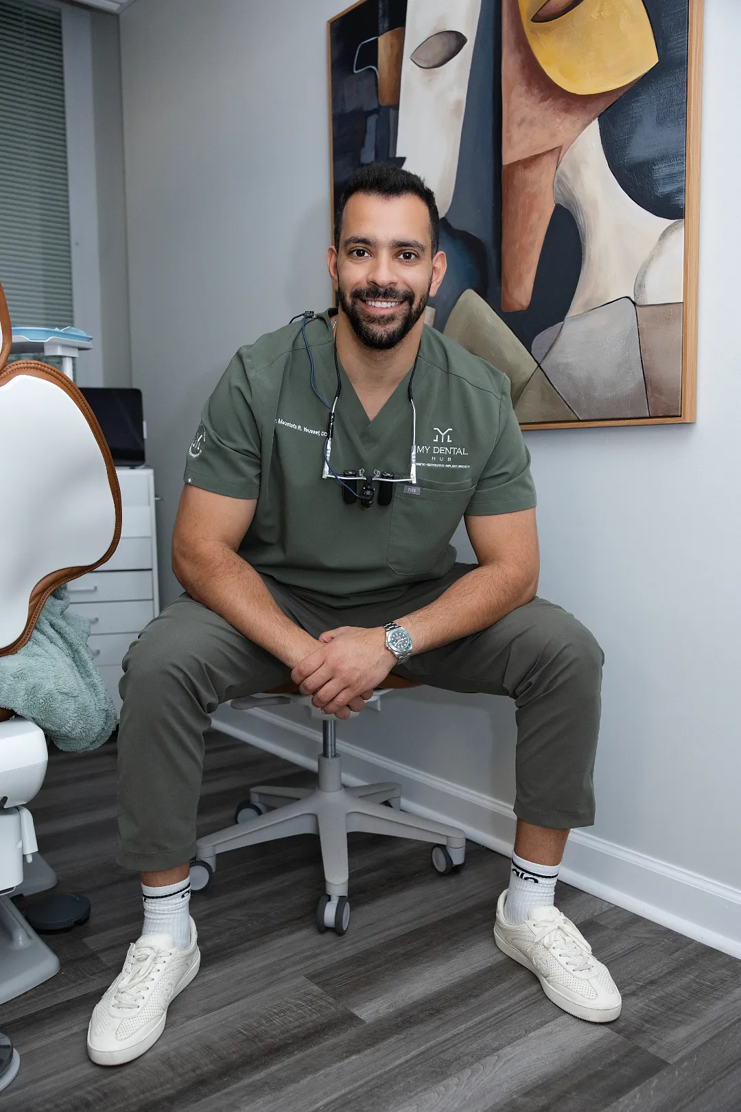 Smiling adult male dentist in green scrubs sitting on a rolling stool in an operatory — My Dental Hub in Skokie, IL
