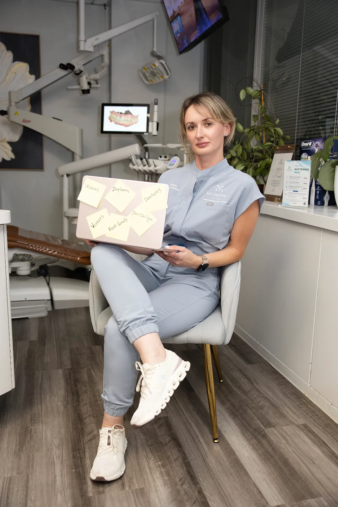 Female dentist in grey scrubs holding a laptop covered in sticky notes listing dental services in an operatory — My Dental Hub in Skokie, IL