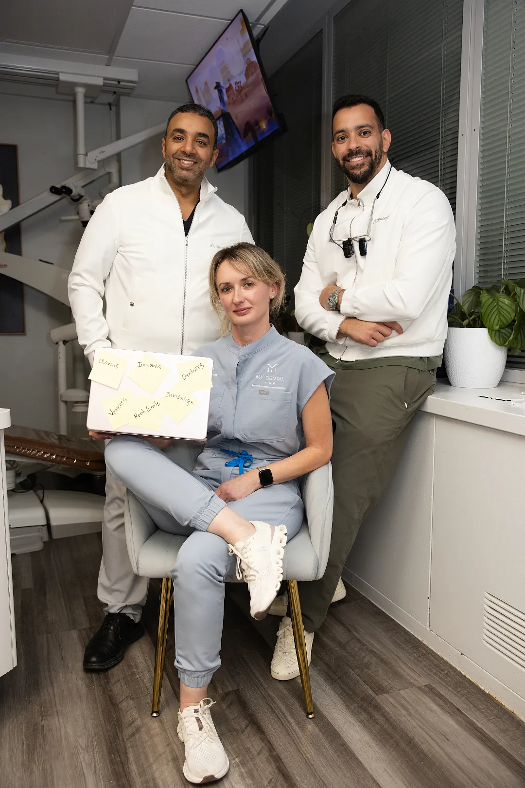 Three dental staff members posing in a clinic, one seated holding a laptop with sticky notes — My Dental Hub in Skokie, IL